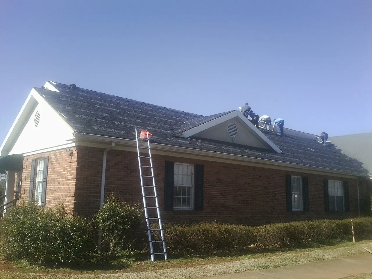 Crew tearing shingles off a brick ranch roof with a ladder set — Emergency Roof Repair work by our crew in Mc Calla